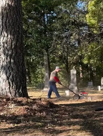 A volunteer pushes a mower among trees and grave markers.
