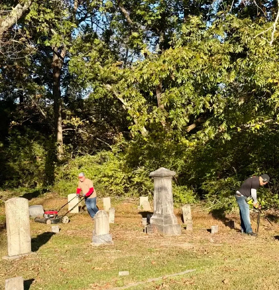 Two volunteers mow and trim vegetation among cemetery grave markers.