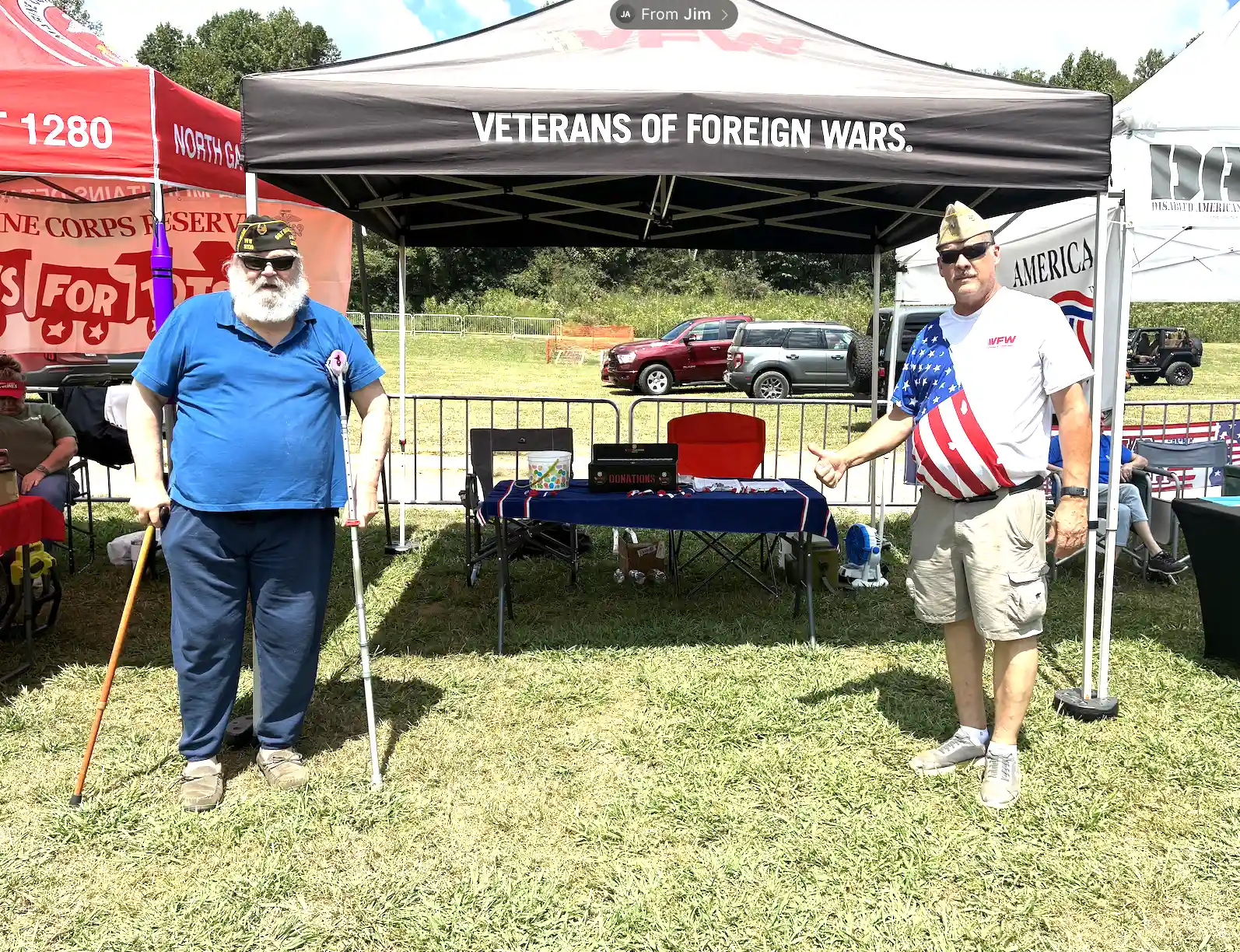 Two volunteers stand beside the Buddy Poppy display and donation box under the VFW canopy.