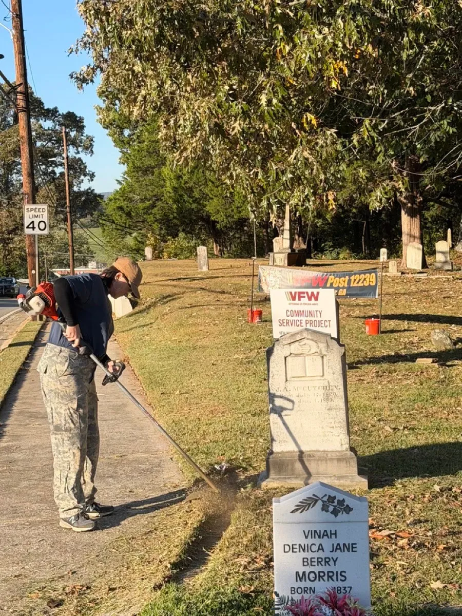 A volunteer uses a string trimmer along the sidewalk beside cemetery grave markers.